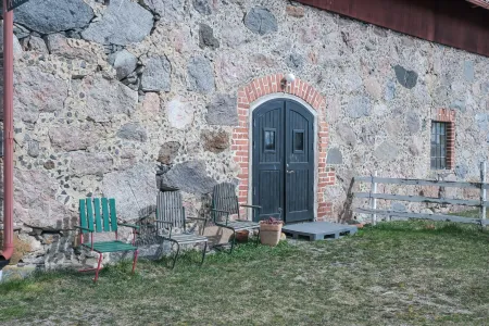 En rustik stenvägg med mörkgrön dubbeldörr i tegelvalv och tre tomma stolar på gles gräsmatta. (illustrationsbild)