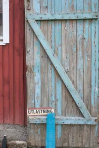 En väderbiten trädörr med flagnande ljusblå färg och skylten UTLASTNING mot röd träfasad.