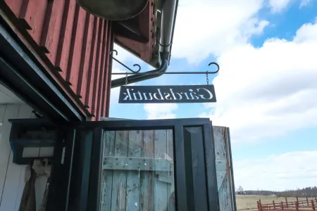 En svart smidesskylt med texten Gårdsbutik hänger på en faluröd träbyggnad under klarblå himmel. (illustrationsbild)