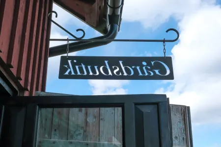 En svart smidesskylt med utskuren text Gårdsbutik ses bakifrån mot röd träfasad och blå himmel. (illustrationsbild)