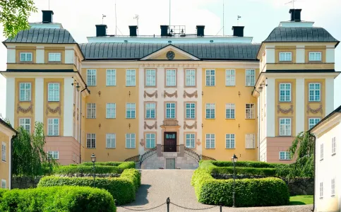 Hela huvudfasaden på Eriksbergs slott med flyglar, klippta häckar och grusgång under klar sommarhimmel.