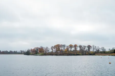 En lugn vattenvy mot en trädbevuxen udde i gula och orangea höstfärger under mulen himmel.