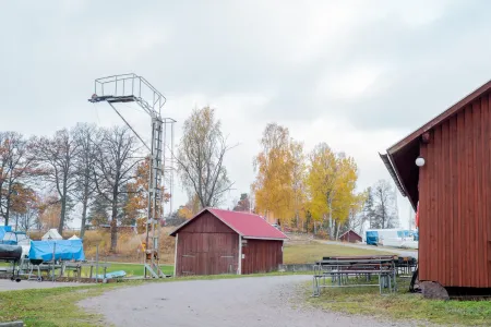 Röda träbyggnader och en båtkran vid en grusväg i ett hamnområde med gula höstträd. (illustrationsbild)