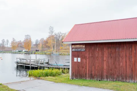 En rödmålad träbyggnad med skyltar Minilivs och Gatubod står vid brygga och båtramp i höstlandskap.