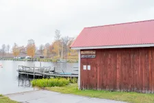 En rödmålad träbyggnad med skyltar Minilivs och Gatubod står vid brygga och båtramp i höstlandskap.