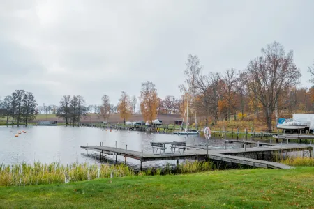 En träbrygga med bänkar sträcker sig ut i vattnet vid en båtplats omgiven av höstfärgade träd.