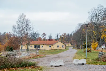 En gul träbyggnad med rött tegeltak omges av höstfärgade träd och en grusväg med betonghinder.