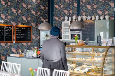 En kund i grå kappa och blå mössa står vid en cafédisk med bakverk framför blommig tapet. (illustrationsbild)