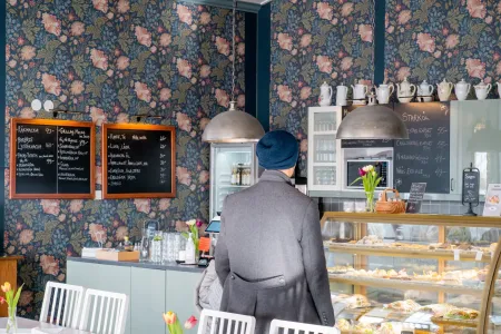 En person står framför en välfylld monter med bakverk och smörgåsar i ett café med blomtapet. (illustrationsbild)