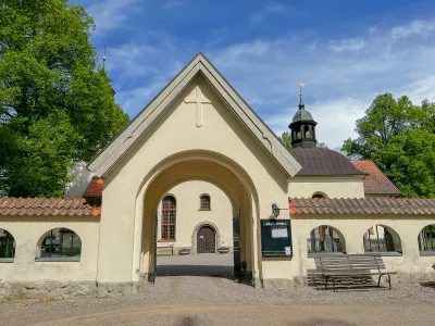 En ljus stenportal med kors och öppna järngrindar leder in till Julita kyrka under klarblå himmel.