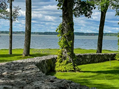 En låg stenmur och lummiga träd i en park med picknickbord och utsikt över en sjö.