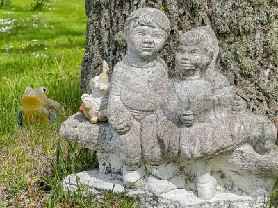 En väderbiten stenstaty av två barn på en bänk står framför en trädstam i blommande gräs.