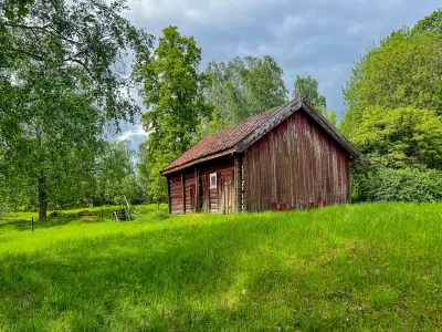 En väderbiten röd träbyggnad med tegeltak står i högt grönt gräs bland lummiga träd.