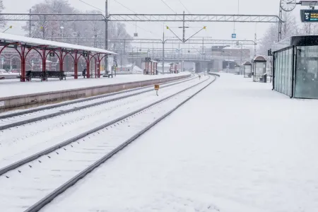 Snötäckta järnvägsspår och perrong vid Katrineholms station med röd väderskärm och elektrifierade kontaktledningar.