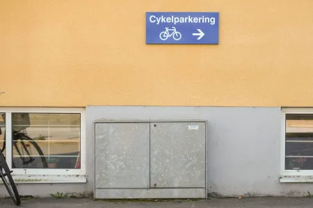 En blå skylt med texten Cykelparkering och en pil sitter på en gul putsad fasad. (illustration)