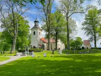 En vit kyrka med klocktorn och tegeltak omges av gravstenar och grönskande lövträd.