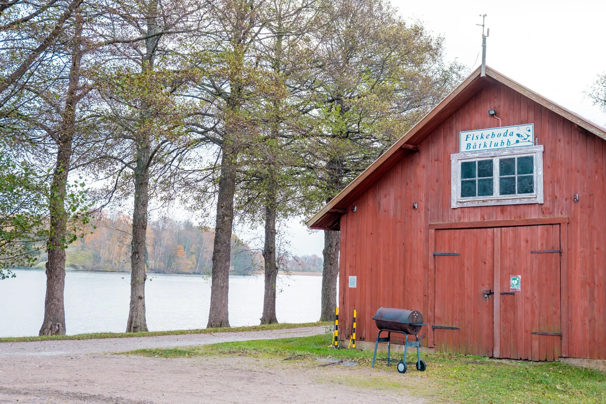 Ett rött trähus med skylten Fiskeboda Båtklubb står vid sjön omgiven av höstfärgade träd.