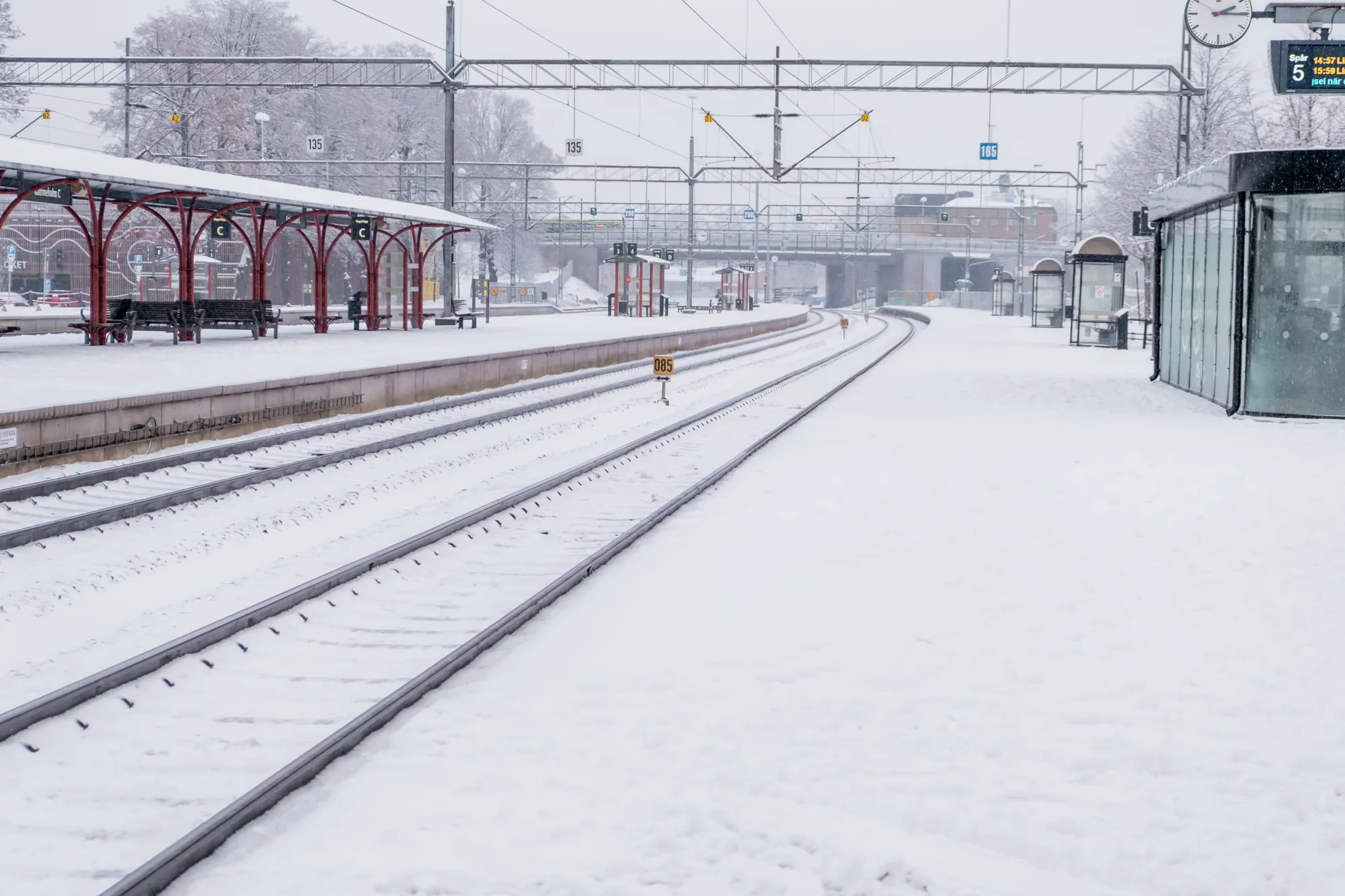 Snötäckta järnvägsspår och perrong vid Katrineholms station med röd väderskärm och elektrifierade kontaktledningar.