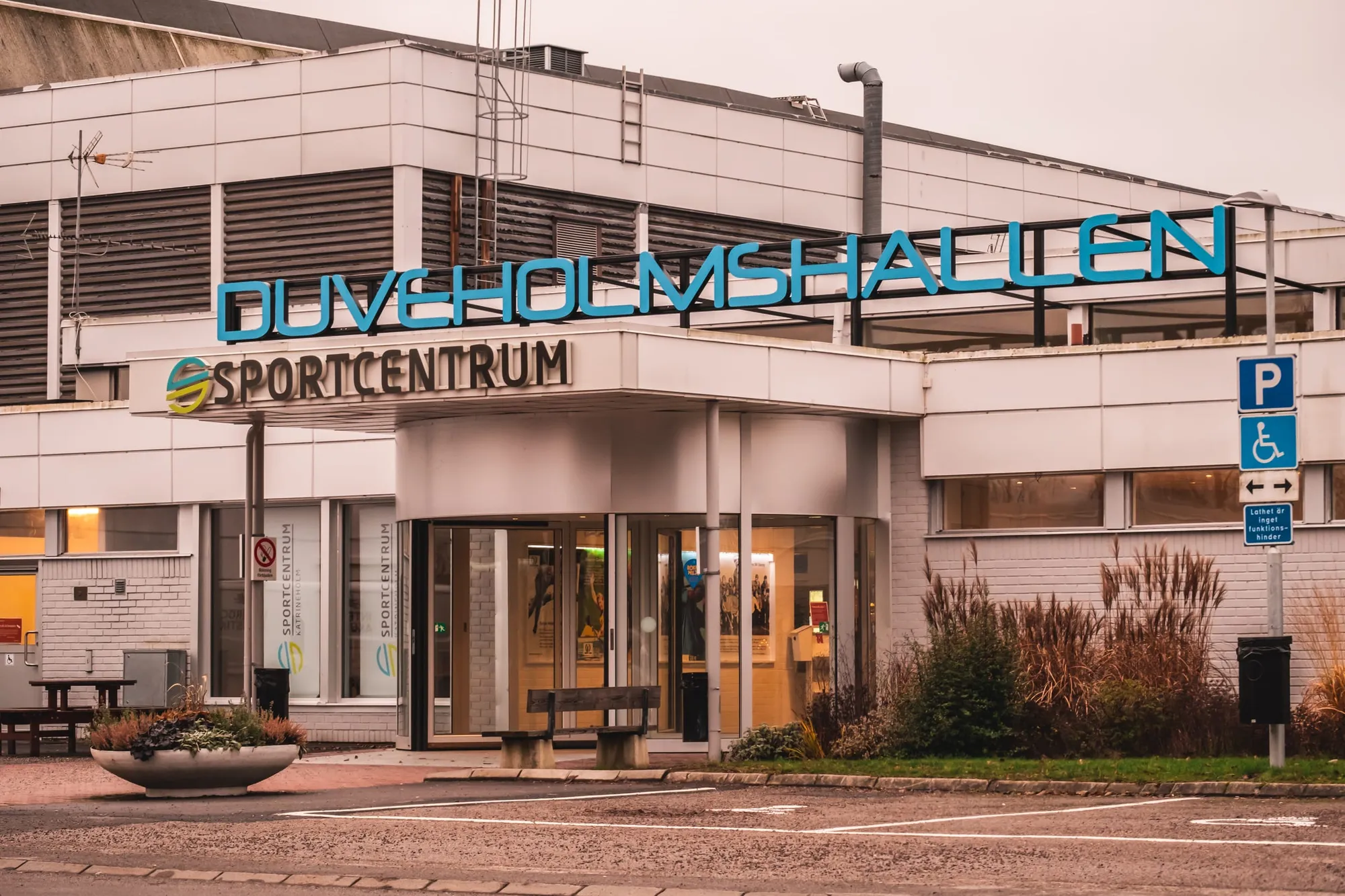 Entrén till Duveholmshallen och Sportcentrum syns från gatan med parkering i förgrunden.